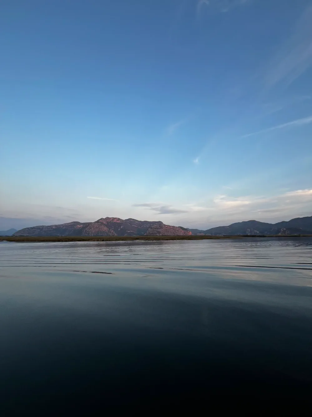 Dalyan deltasında geniş ve durgun su yüzeyi; karşıda uzayıp giden dağ sırası ve ince sazlık şeridi