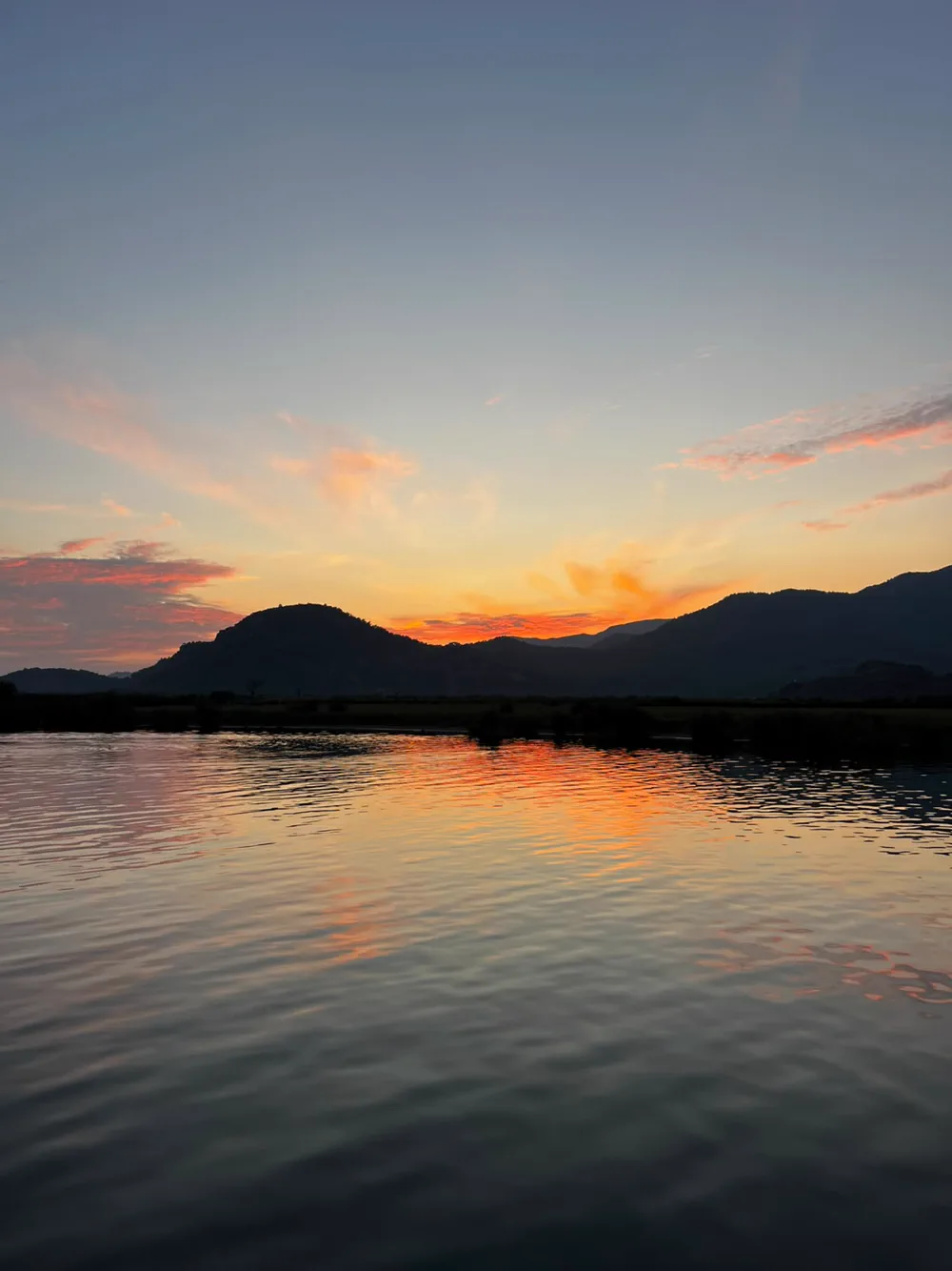 Dalyan Çayı üzerinden gün batımı: turuncuya dönmüş gökyüzü, karşıda dağ siluetleri ve suya düşen uzun yansıma