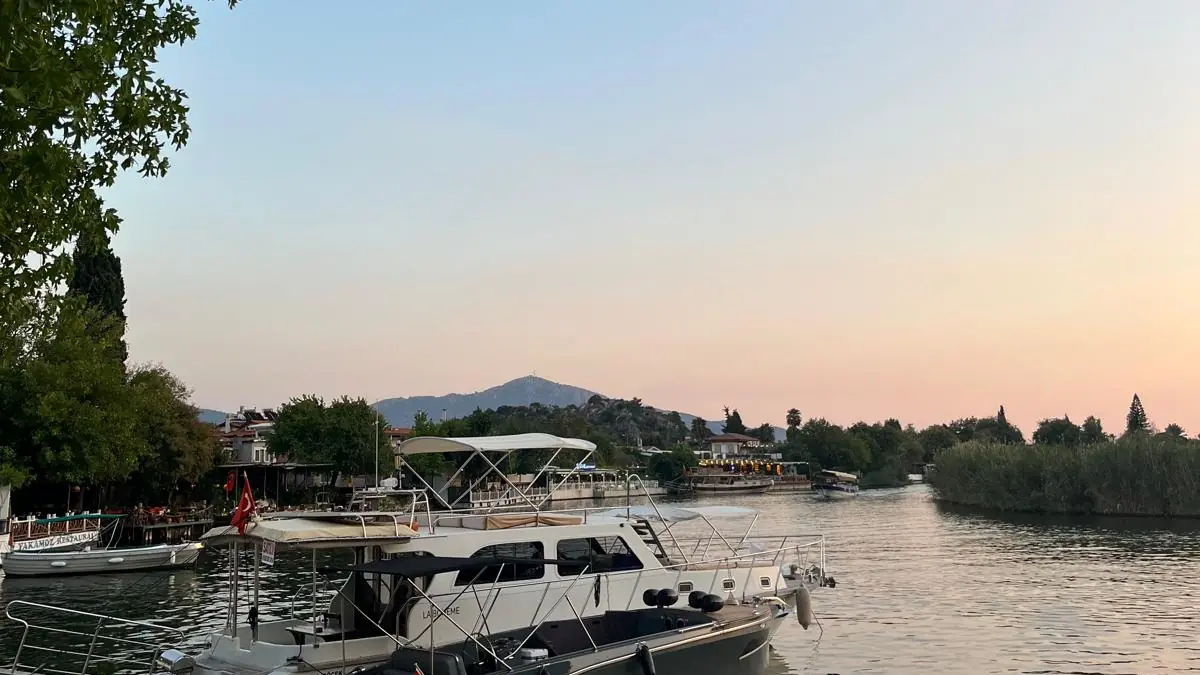 Akşamüstü Dalyan iskelesine bağlı tur tekneleri; nehir ilerideki dağlara doğru uzanıyor, gökyüzü turuncuya dönmüş