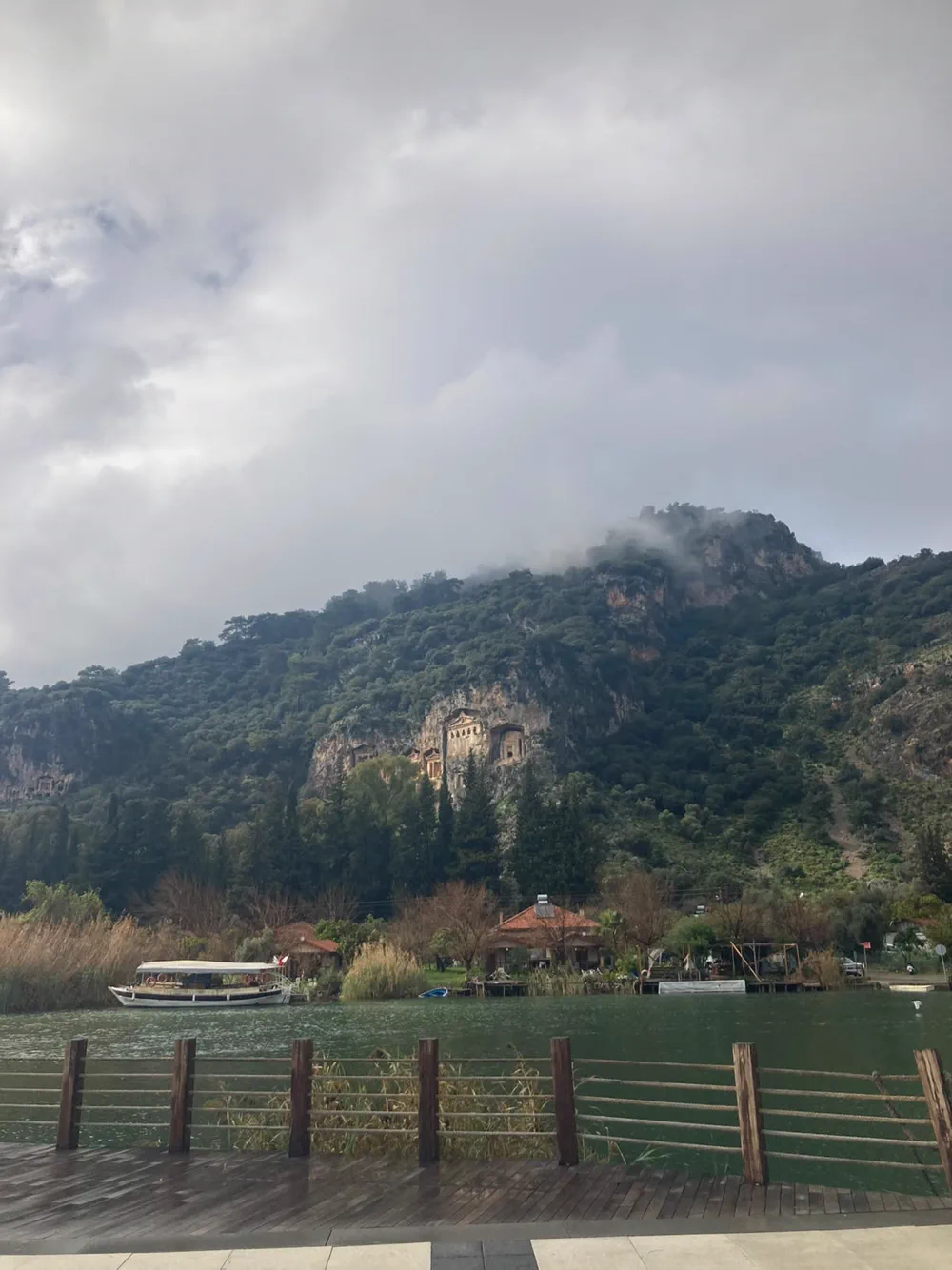 Bulutun kayalığa indiği bir günde Dalyan kaya mezarları; suda bağlı bekleyen bir tekne ve ahşap iskele korkuluğu