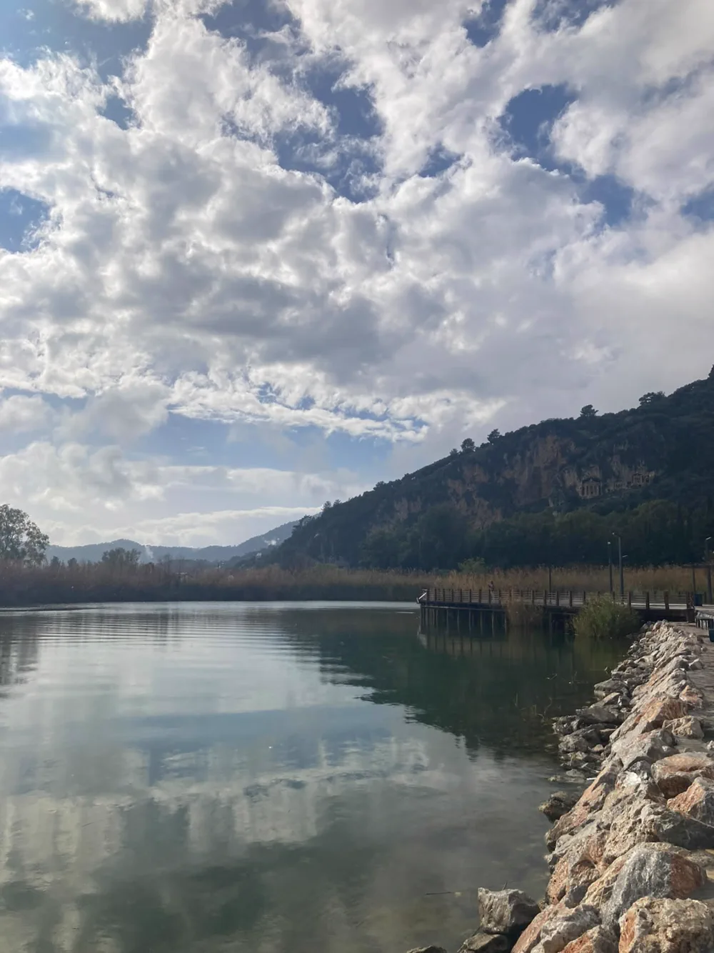 Dalyan kordonunun kıyı taşları, ahşap iskelesi ve karşı kayalıktaki mezarlar; su ayna gibi duruyor