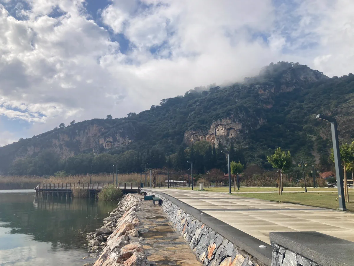 Dalyan kordonunun taş döşeli yürüyüş yolu, kıyıdaki ahşap iskele ve karşı yamaçta Kaunos nekropolündeki tapınak cepheli kaya mezarları