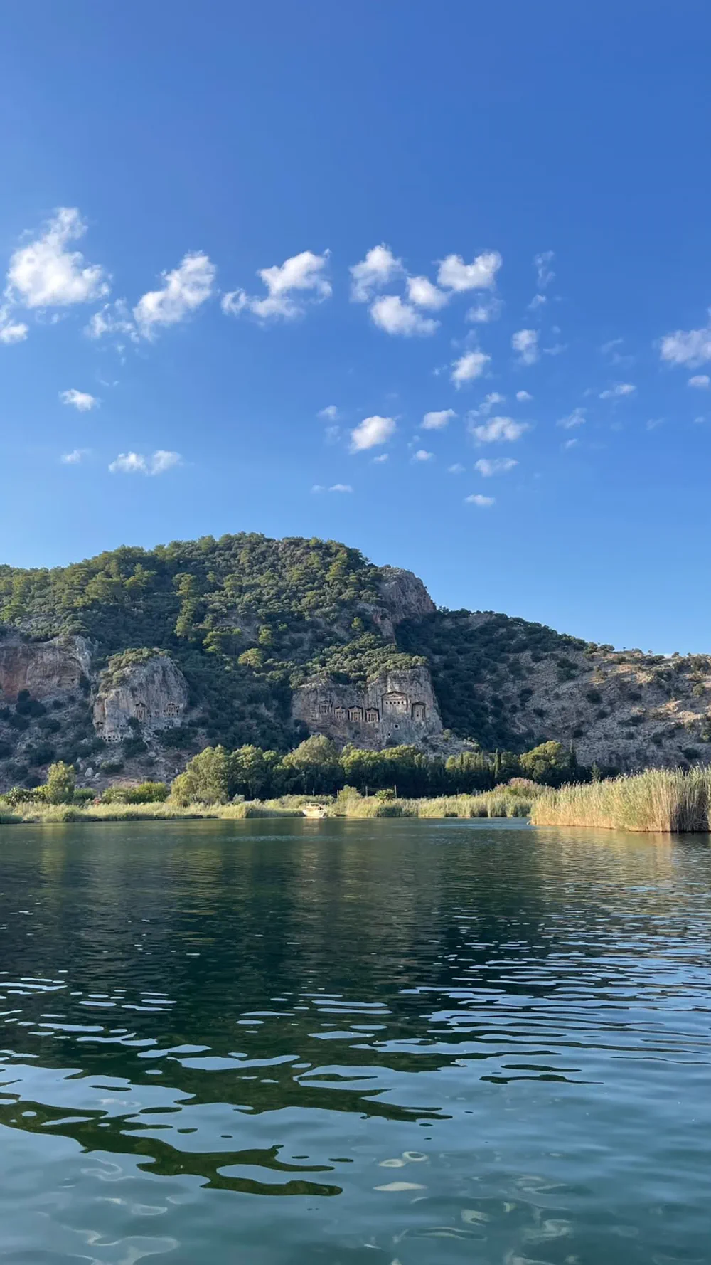 Yaz günü sudan Dalyan kaya mezarları: sazlık şeridi, karşıda bağlı bir tekne ve berrak nehir