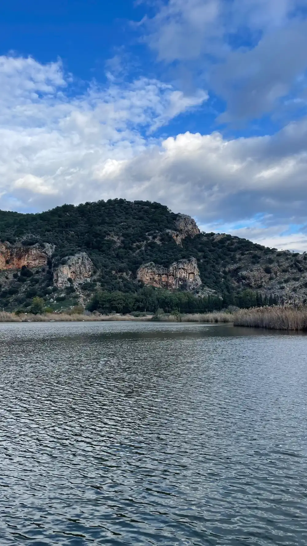 Bulutlu bir günde Dalyan Çayı ve karşı yamaçtaki kaya mezarları; kıyıda sararmış sazlıklar