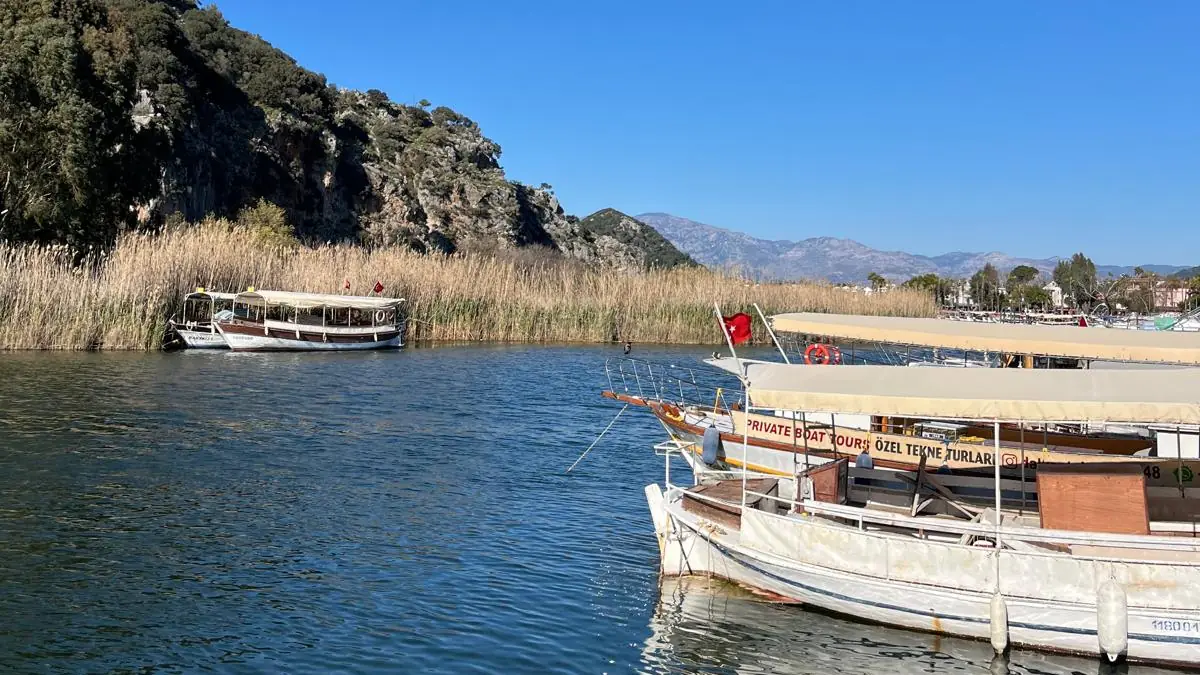 Dalyan Çayı kıyısına bağlı, üzerinde özel tekne turları yazan geleneksel ahşap tur tekneleri; arkada sazlıklar ve kayalık yamaç