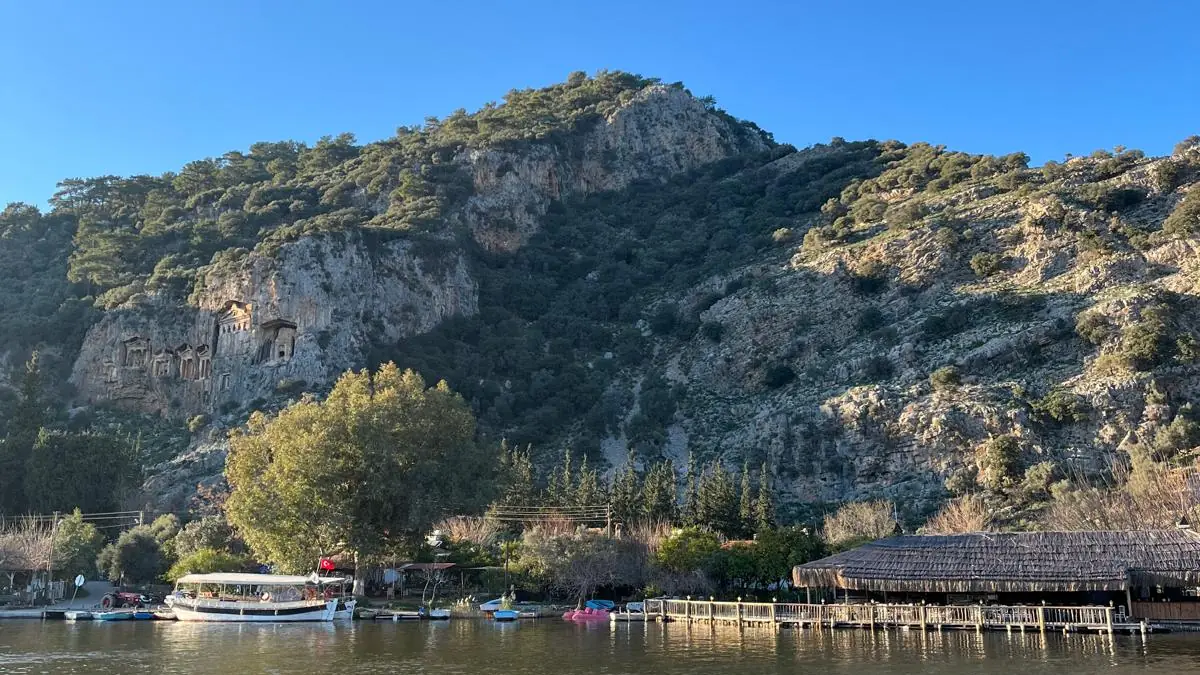 Dalyan Çayı’nın karşı kıyısındaki Kaunos kayalığı; yamacın solunda kaya mezarları, altında kordondaki tur tekneleri ve iskele